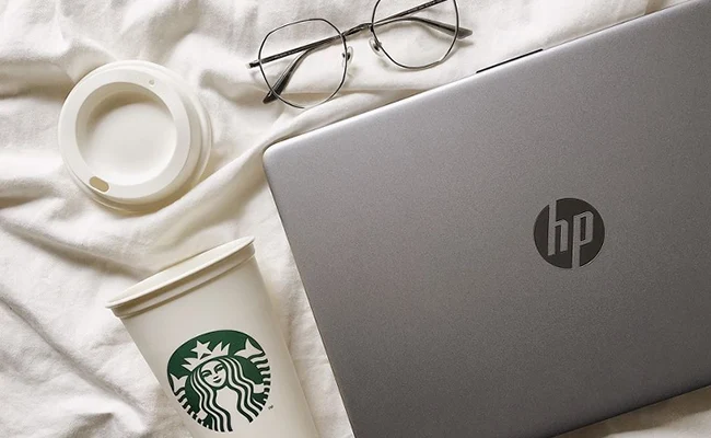 عکس لپ تاپ hp روی تخت کنار عینک و لیوان استارباکس